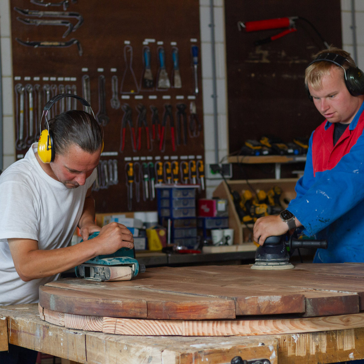 Twee mannen met gehoorbescherming schuren samen een groot rond houten werkstuk in een goed uitgeruste werkplaats.