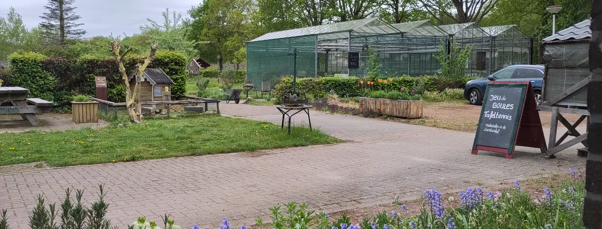 Erf boerderij De Stiep met bloeiende planten, wandelpaden en een grote kas, met een krijtbord bij de ingang.