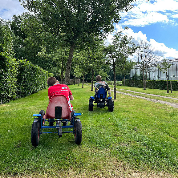 Twee jongens rijden op skelters over een grasveld tussen bomen, met een kas op de achtergrond.